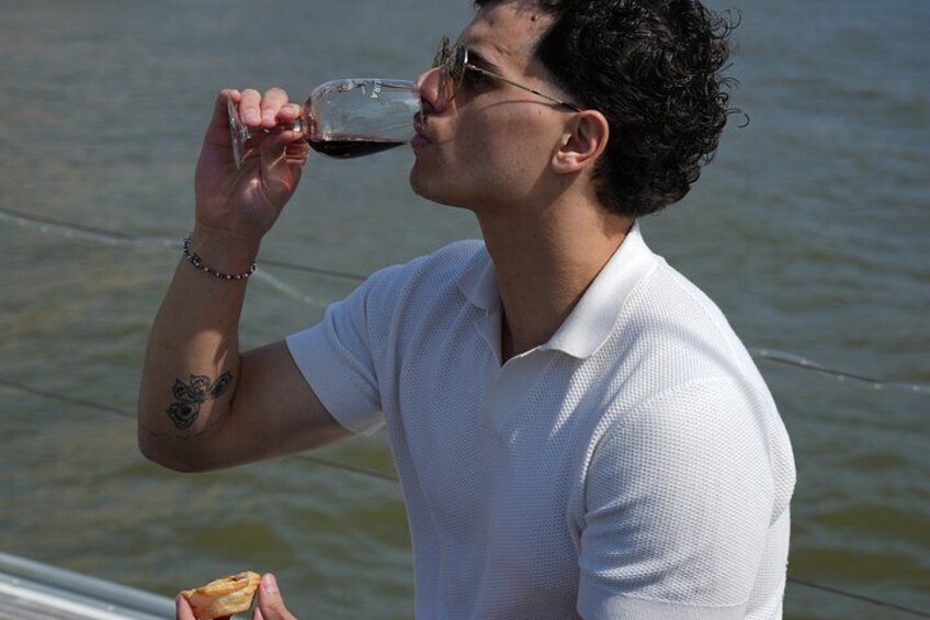 Iemand drinkt uit een glas rode wijn, draagt een wit shirt en heeft een tatoeage op hun arm, staand bij een waterpartij. 3