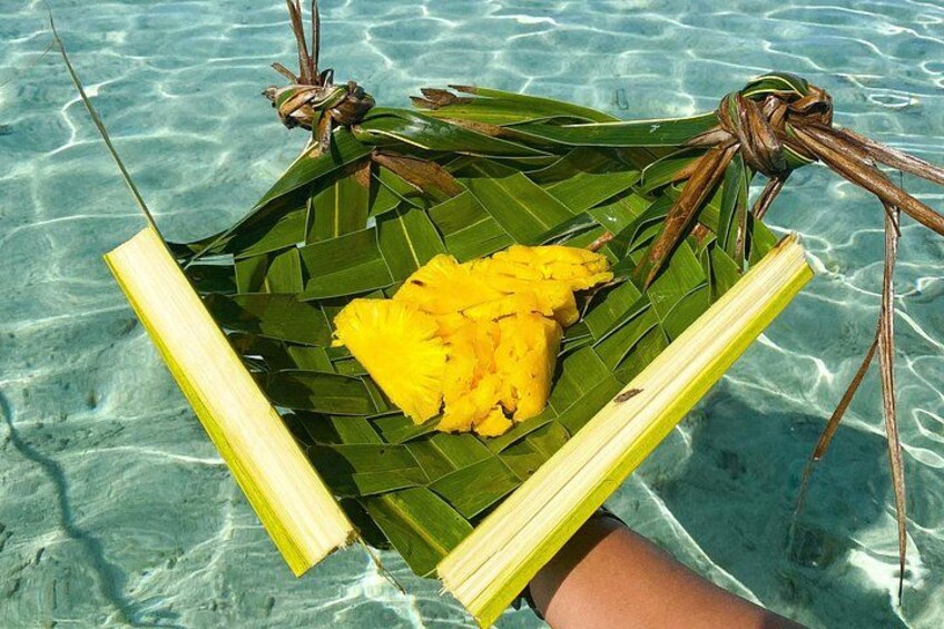 A person holding a woven basket with a yellow fruit and green leaves over clear blue water. 2