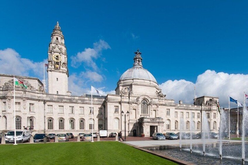 A large historic building with a clock tower and a fountain in front. 1