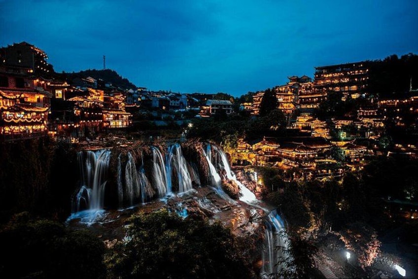 A nighttime waterfall cascading down a rocky cliff surrounded by illuminated buildings. 4