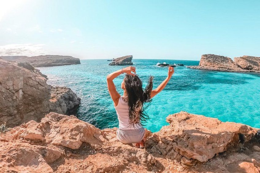A person sitting on rocks by a clear blue sea with rocky cliffs in the background. 2