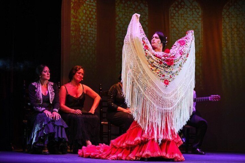 A stage with three people, one in a red dress with a fringe shawl, and two others in dark attire. 3