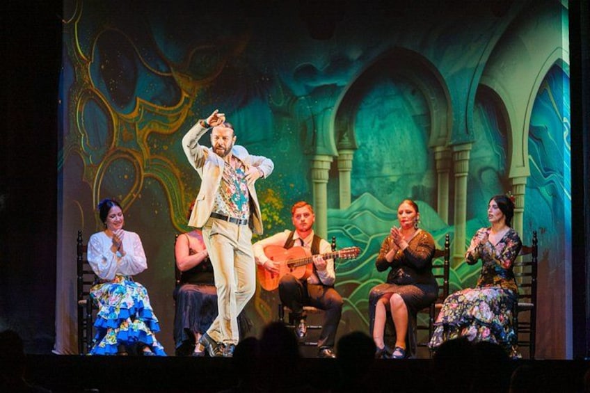A stage with five people, one standing and four seated, with a backdrop of arches and abstract designs. 3