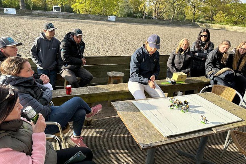 A group of people sitting outdoors on benches and chairs, with a table in the foreground that has a chessboard and some figurines. 3