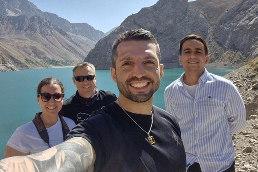 Eine Gruppe von Personen macht ein Selfie am See, mit Bergen im Hintergrund. 1