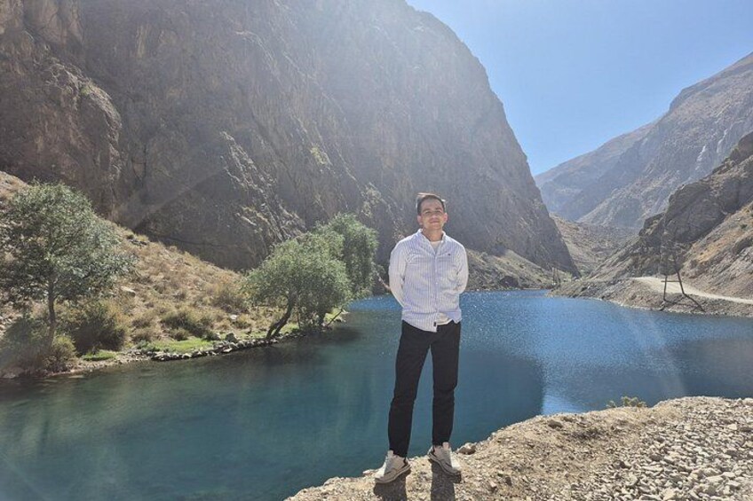 Una persona de pie junto a un río tranquilo en una zona montañosa con terreno rocoso y vegetación escasa. 2