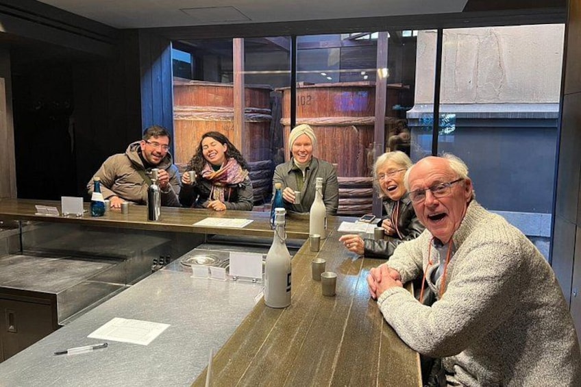 A group of people seated at a table with a bottle and glasses, in a room with a large window and wooden elements. 2