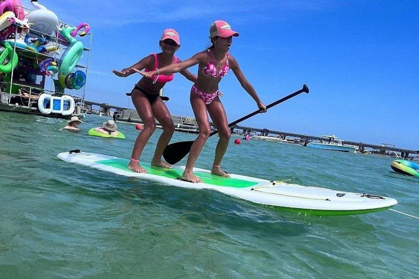 Zwei Personen stehen auf Paddleboards auf ruhiger See, im Hintergrund ein Steg und aufblasbare Boote. 3