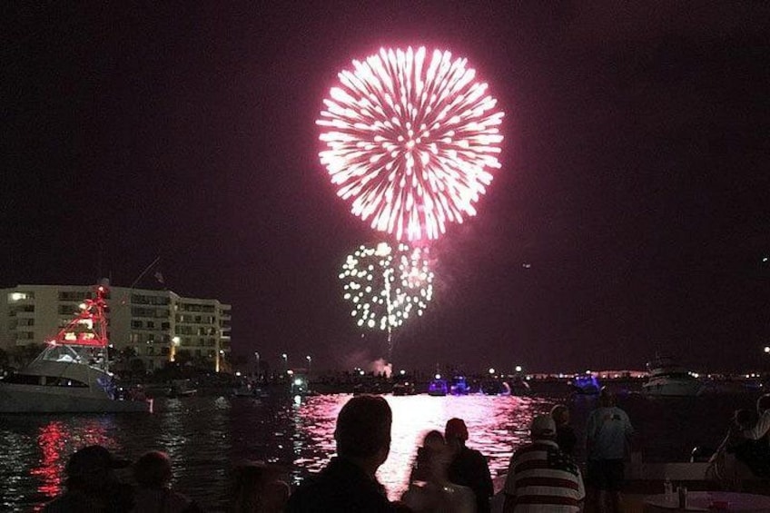 Escena nocturna con fuegos artificiales sobre el agua, edificios al fondo y gente en un barco. 3