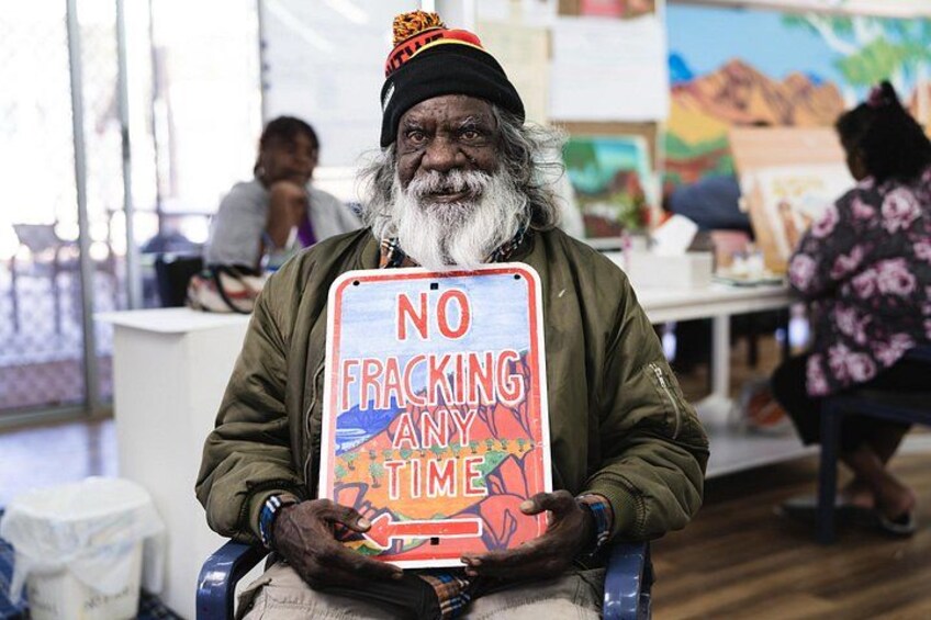 A person holding a sign that reads 'NO FRACKING ANYTIME' in a room with tables and chairs. 4