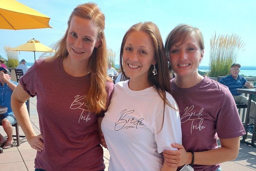 Tre personer iført 'Bride Tribe'-t-shirts står udendørs med borde og parasoller i baggrunden. 4