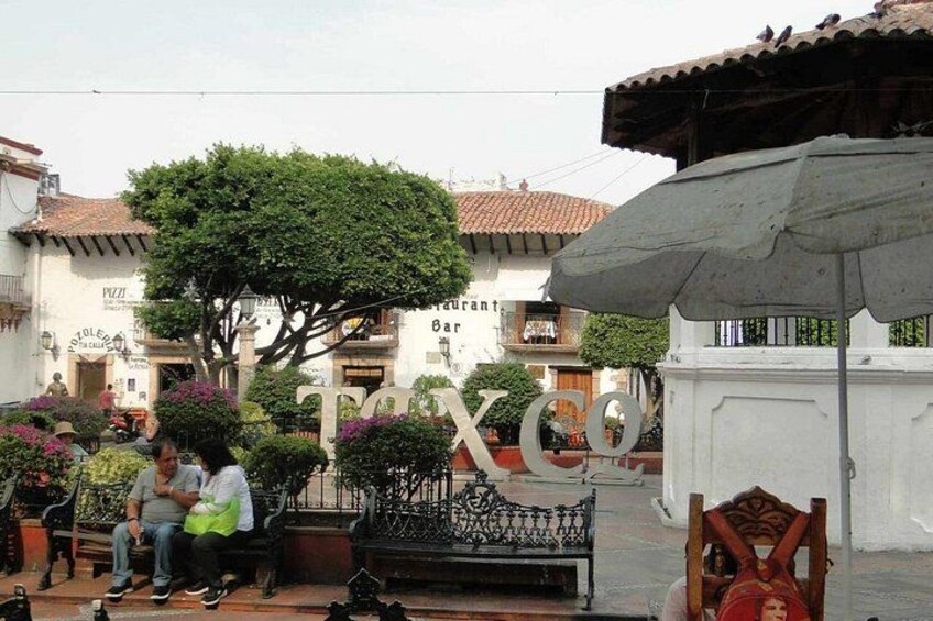 A street scene with a large sign reading 'TAXCO,' people sitting on benches, and a tree in the background. 3