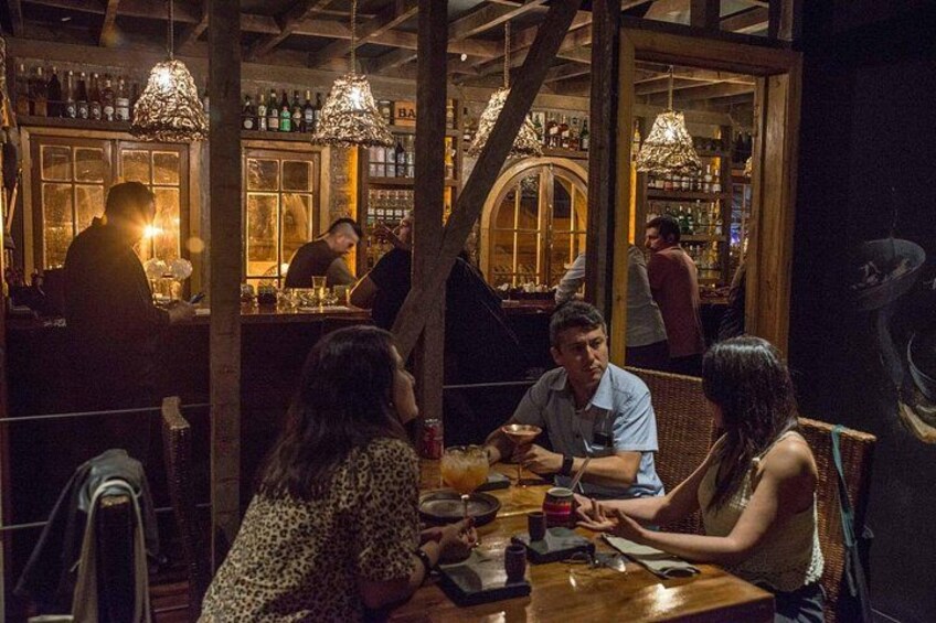Un bar con clientes sentados en mesas, vigas de madera, luces colgantes y estanterías con botellas. 3