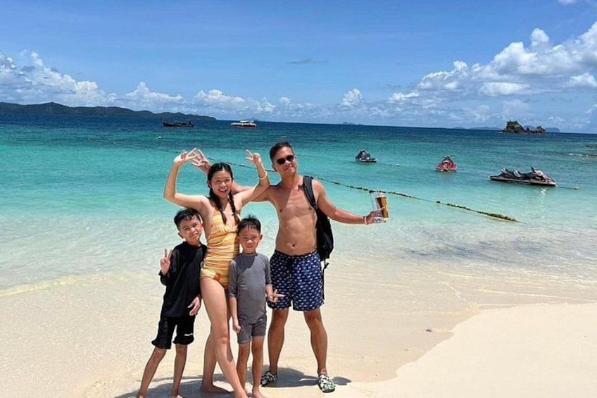 A family on a beach with clear turquoise water, a sandy shore, and boats in the distance. 5