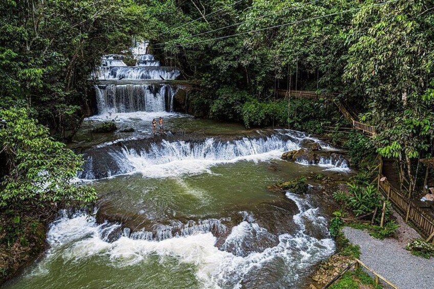 Una cascada fluye sobre rocas en un área boscosa, con una pasarela de madera cerca. 4