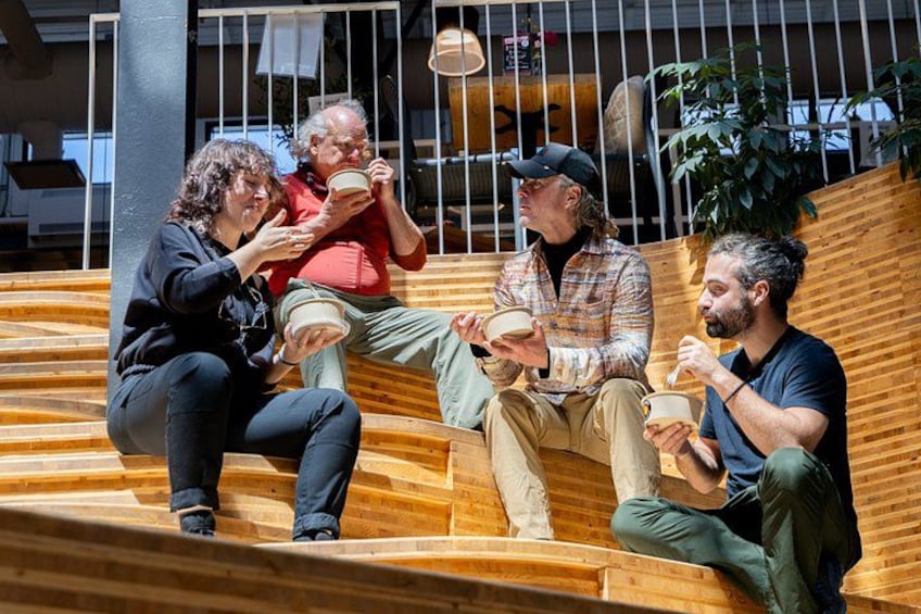 Four people sitting on a wooden bench outdoors, each holding a cup. 2