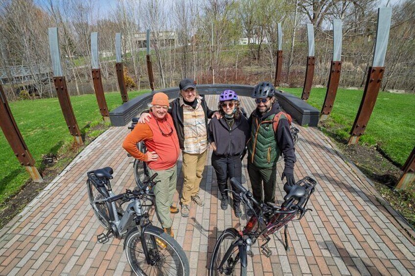 Cuatro personas en un sendero de ladrillo con bicicletas, rodeadas de postes metálicos y follaje. 2