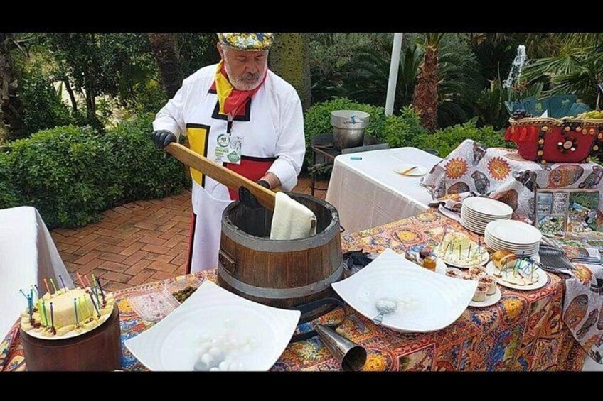Une personne vêtue d’un costume traditionnel prépare de la nourriture à l’extérieur, à côté d’un tonneau en bois, d’une table avec des assiettes et un gâteau, dans une ambiance festive. 4
