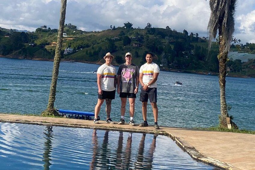 Three people standing by a calm body of water with a wooden dock and palm trees in the background. 2