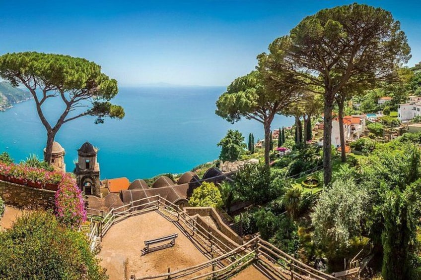 A coastal landscape with a clear view of the sea, a large tree, a small bell tower, and a stone staircase leading down to a bench. 2
