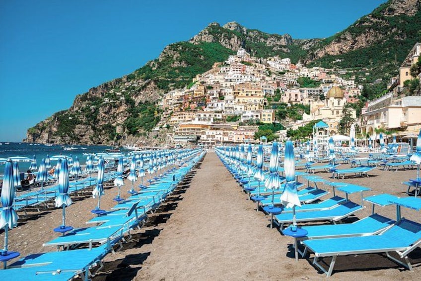 A beach with rows of blue sunbeds and umbrellas, a clear sky, and a hillside town in the background. 4