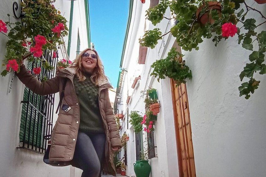 Una persona de pie en una calle estrecha con paredes blancas, contraventanas verdes y flores rojas. 3