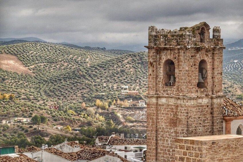 Una torre de piedra histórica con ventanas arqueadas y una campana, con vistas a un paisaje montañoso con vegetación escasa y edificios dispersos. 1