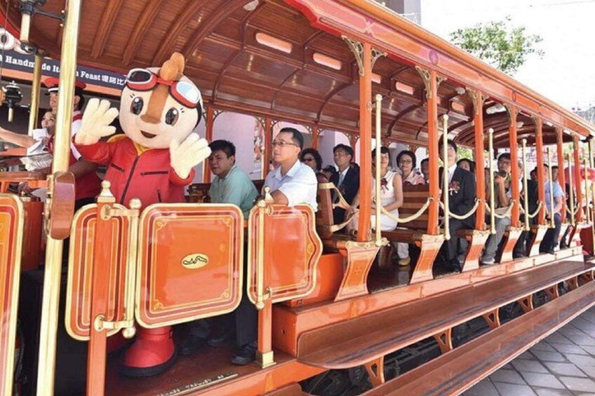 Eine hölzerne Straßenbahn mit Fahrgästen und einer tanzenden Maskotte. 3