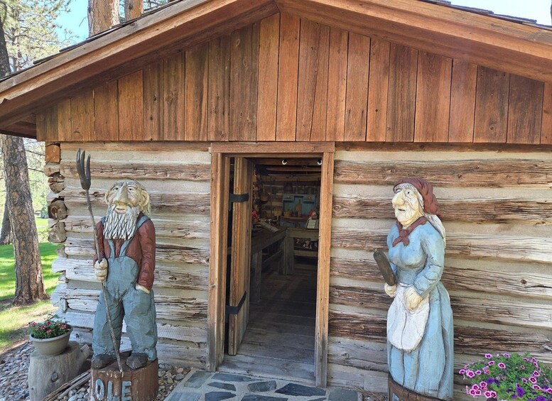 A rustic wooden cabin with a stone foundation, two wooden statues of men, and a flower bed with pink flowers. 4
