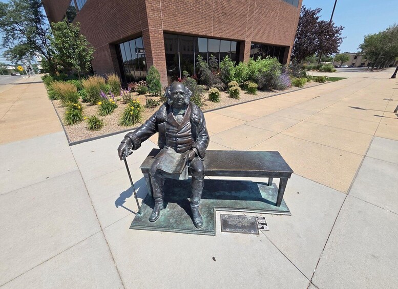 A bronze statue of a seated man with a cane, a bench, and a plaque in front of a building. 5