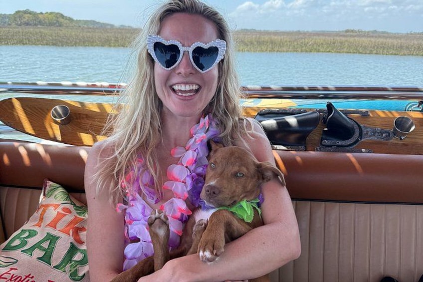 Una persona en un bote con un perro que lleva un collar de flores, rodeado de agua y vegetación. 2