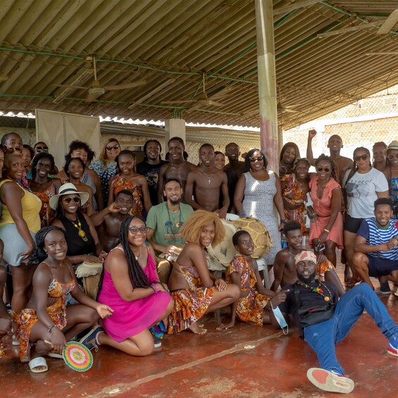 A group of people gathered under a metal roof, some sitting and some standing, in an outdoor setting. 3