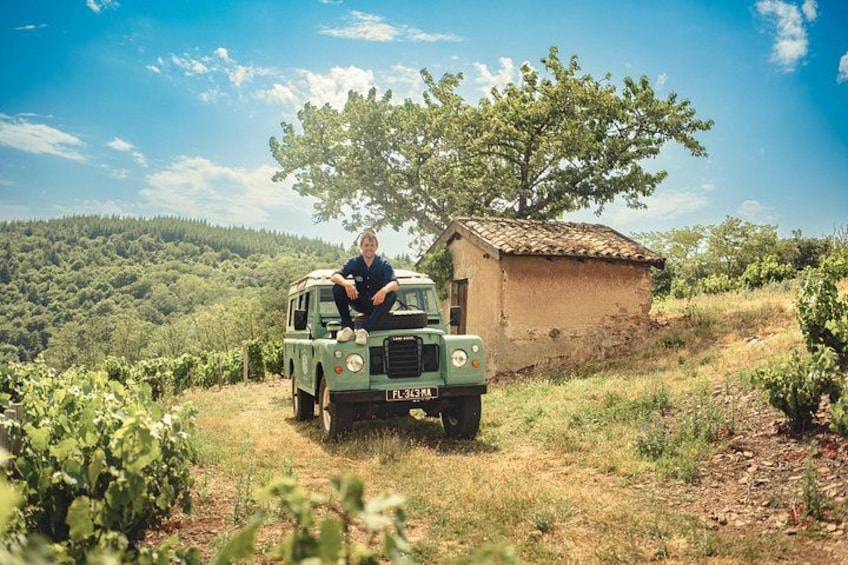 A person sitting in a green vehicle parked near a small building in a hilly, vineyard area. 1