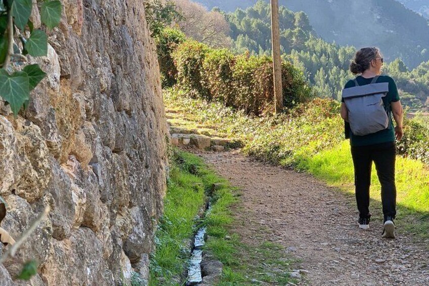 Une personne marchant sur un sentier de terre, avec un sac à dos, entourée de verdure et d’un mur de pierres. 3