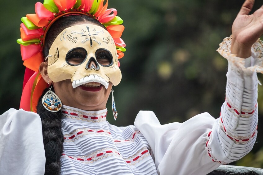 A person wearing a traditional white dress with red and gold embroidery, a colorful floral headdress, and a skull mask. 1