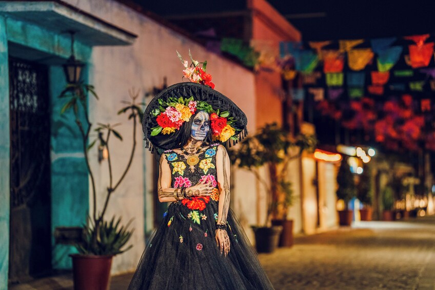 A person in a black dress with colorful floral patterns and a large hat, standing on a cobblestone street at night. 4