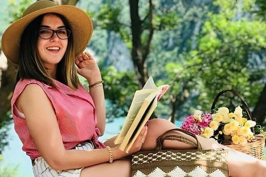 A person sitting outdoors, holding a book, with a basket of flowers and a woven bag nearby. 2