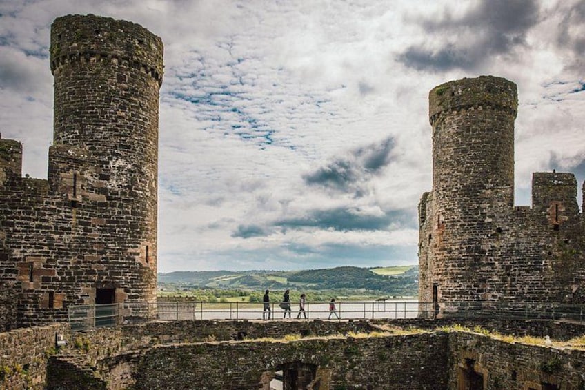 A historic stone castle with two large towers, a paved walkway, and a distant view of green hills under a cloudy sky. 3