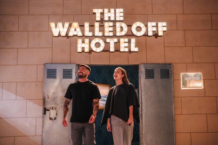 Two people standing in front of a wall with a sign that reads 'THE WALLED OFF HOTEL'. 3