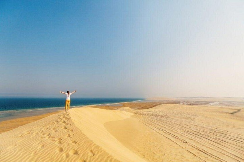 A person standing on a sand dune with arms raised, facing the ocean. 4