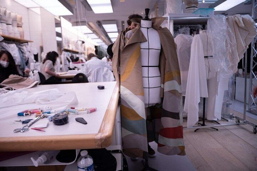 A mannequin draped in a patterned garment, a sewing table with fabric and tools, and a workshop environment with other garments and sewing equipment. 3