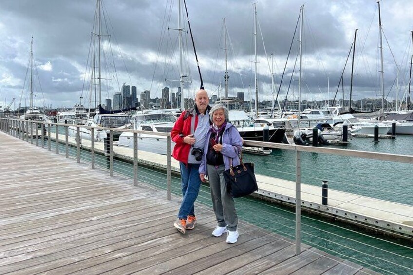 A wooden pier with two people standing, a marina with boats, and a city skyline in the background. 3