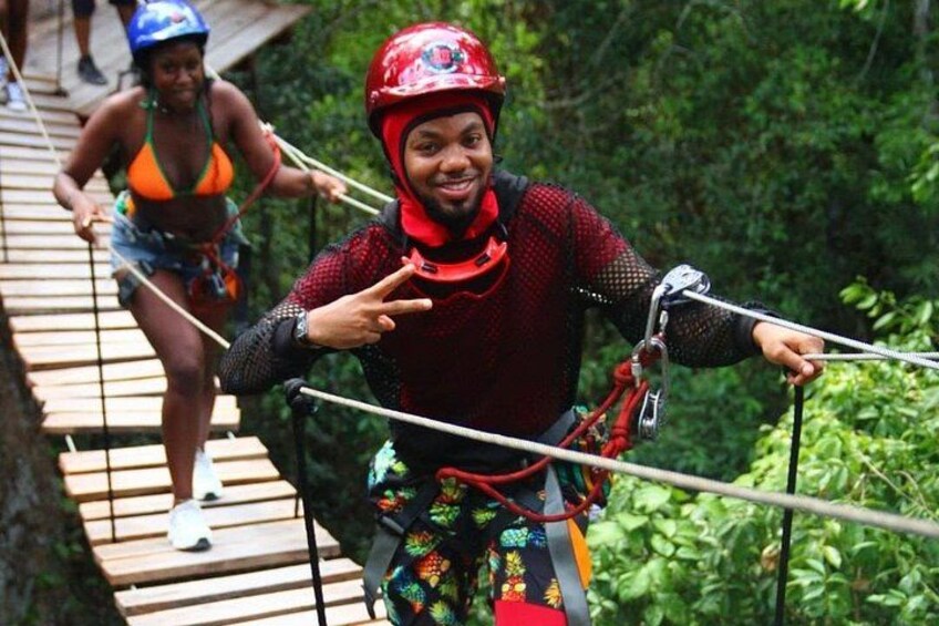 En person på en zipline, utrustad med sele och hjälm, en annan person i bakgrunden på en träbro och ett skogsområde. 4