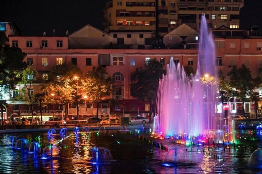 A nighttime cityscape with a brightly lit fountain in the foreground and buildings in the background. 2