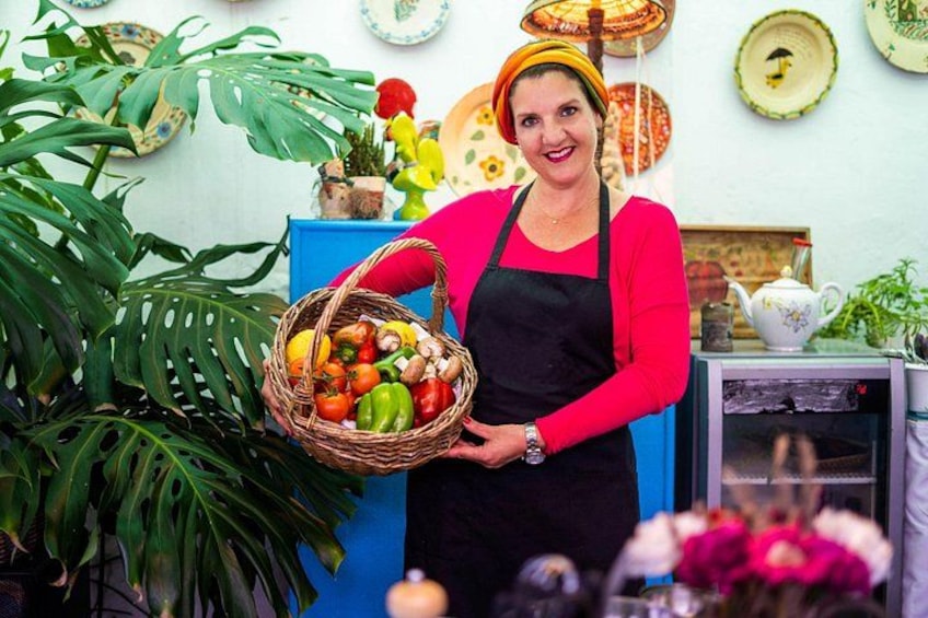 A person holding a basket of vegetables in a kitchen setting with plants and kitchenware in the background. 1