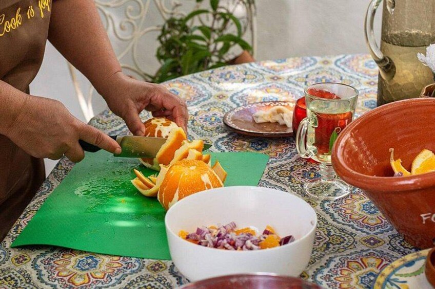 A person peeling an orange on a green cutting board, with bowls of chopped vegetables and a pitcher of water on a patterned tablecloth. 2