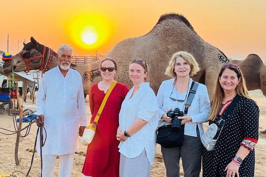 A group of people standing in front of a camel with a desert landscape and a sunset in the background. 1