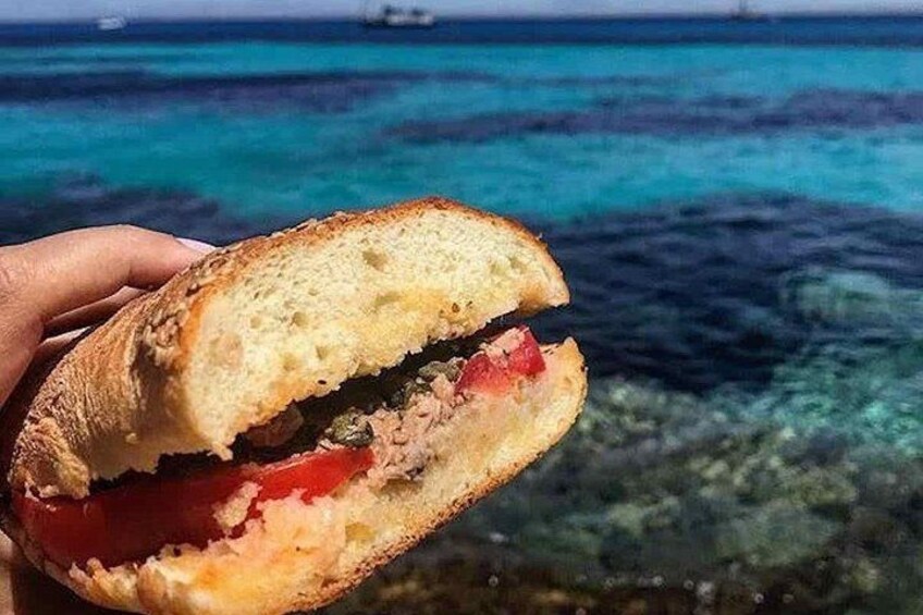 Un sandwich composé de deux tranches de pain, coupées en deux, révélant une garniture de viande et de légumes, le tout mis en valeur par un arrière-plan d’eau d’un bleu limpide et un bateau au loin. 1