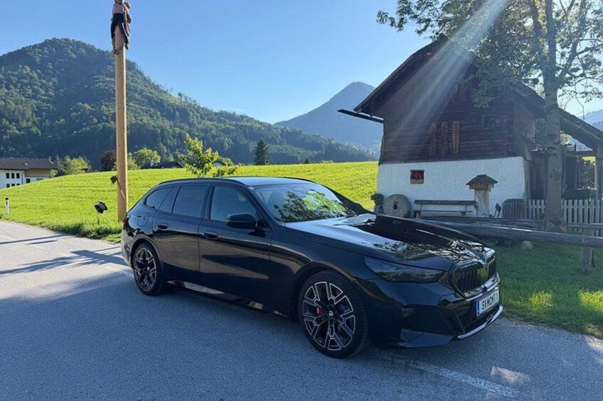 Ein schwarzes Auto steht an einer Straße, im Hintergrund ein Berg und ein Holzhäuschen. 2