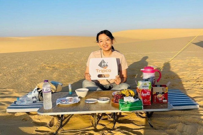 A person sitting on a blanket in a desert with a table of food and drinks. 5